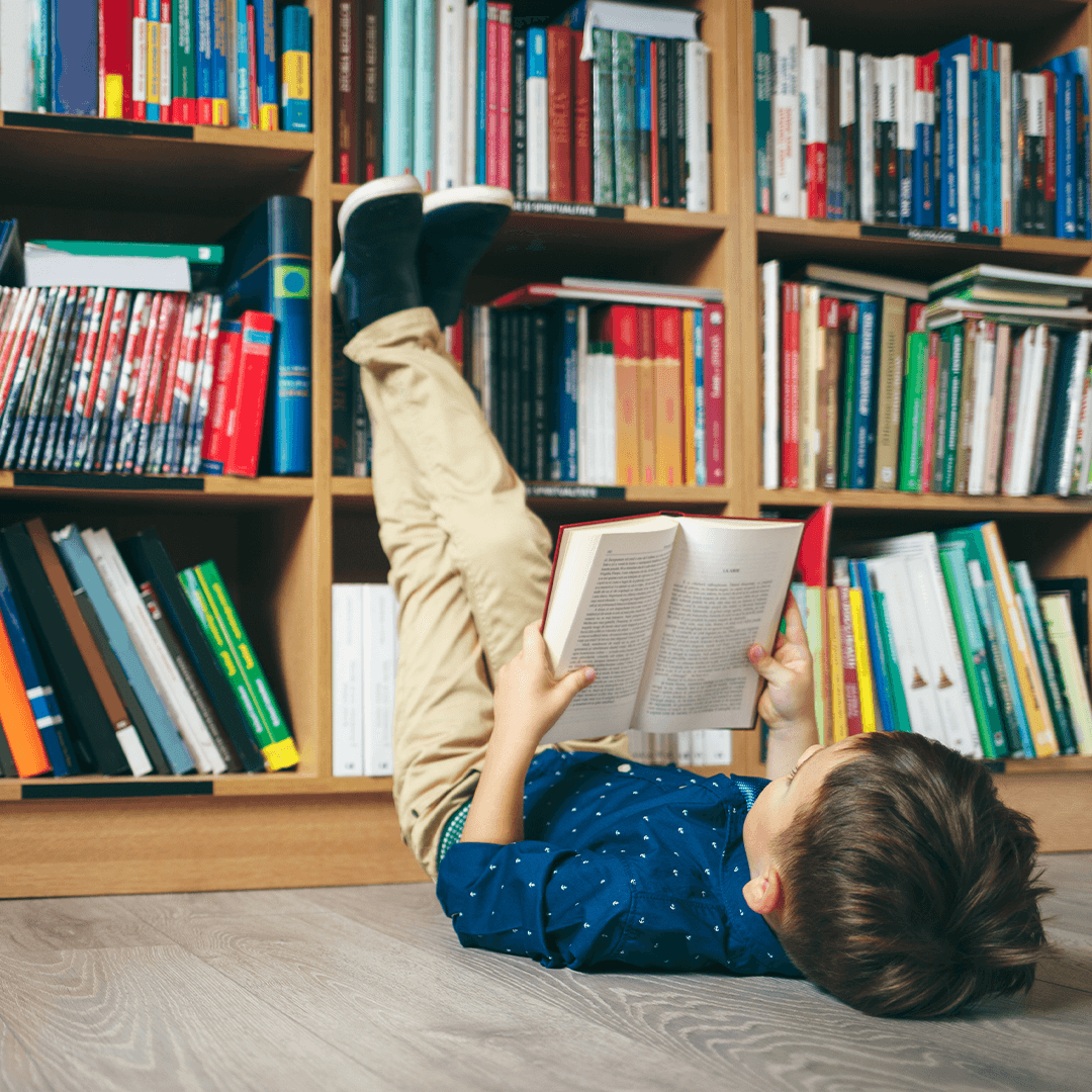 Boy lying on his back reading a book in the library – relaxed moment of imagination