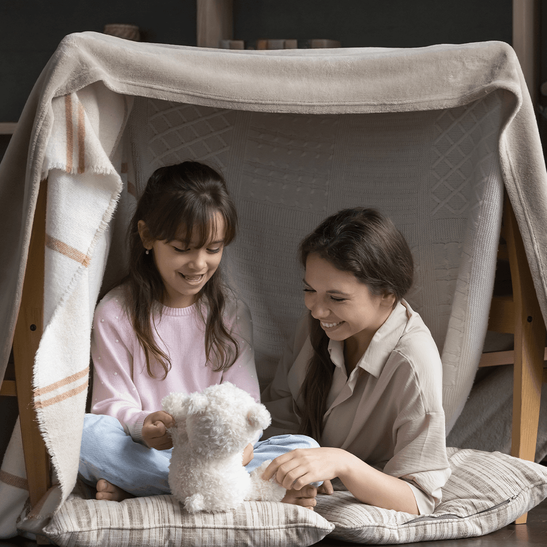 Äiti ja tytär leikkivät viltistä ja tuoleista tehdyssä majassa – leikki-ideoita lapsille sadepäivään.